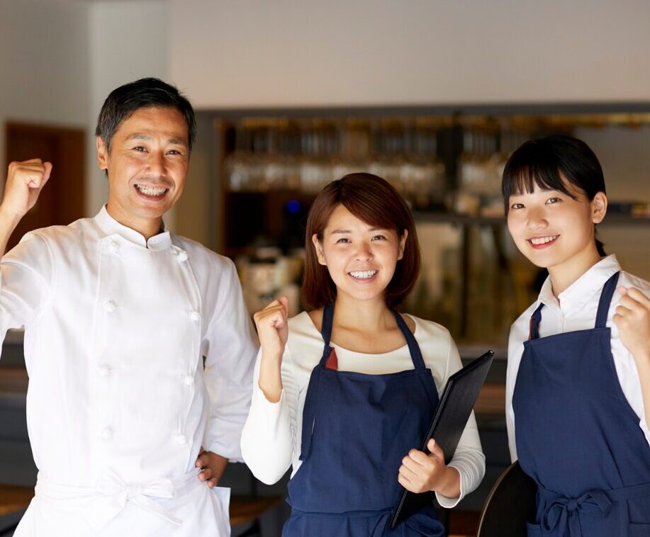 飲食店でガッツポーズをするコック服の男性とエプロン姿のスタッフ2名